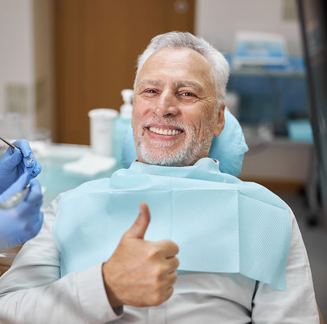 Senior man in the dental chair giving a thumbs up and smiling with implant dentures in Sparks